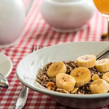 A healthy breakfast bowl with sliced bananas and cereal on a red checkered tablecloth.