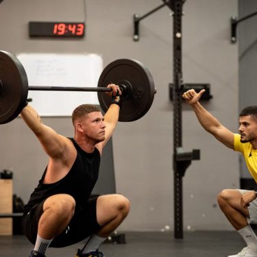 Athlete performing weightlifting exercise under trainer's guidance in a gym setting.